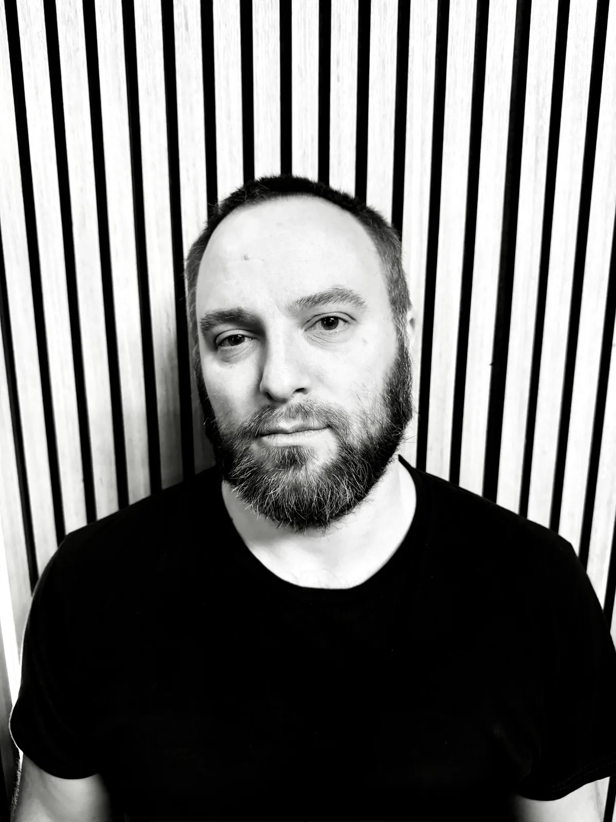 Andrew, with a dark beard and black tee, in front of a slatted wall