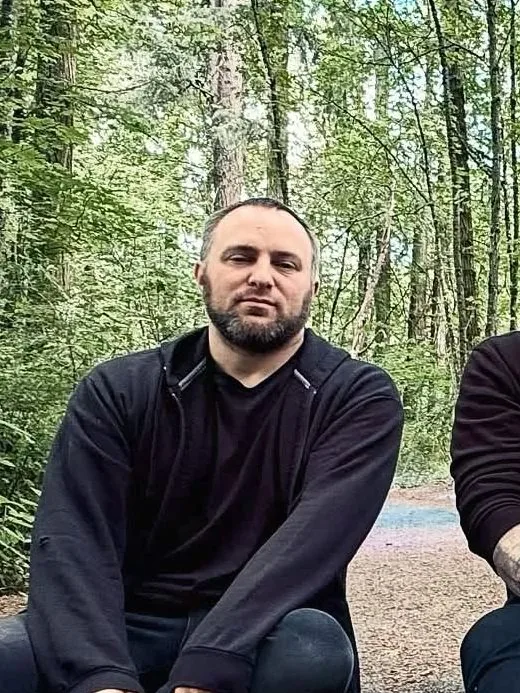 Andrew, with a dark beard and black zip hoodie, sitting in the forest