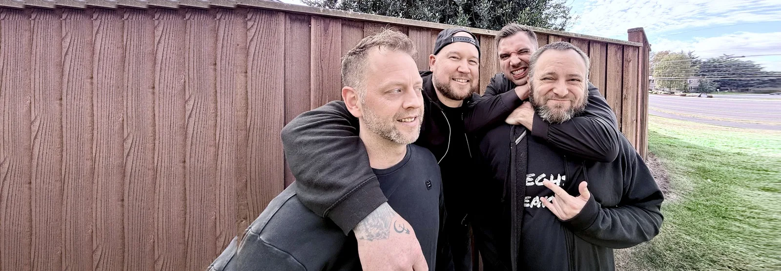 Michael, Graham, Kramer and Andrew of CARD3N arm in arm and grinning in front of a wooden fence