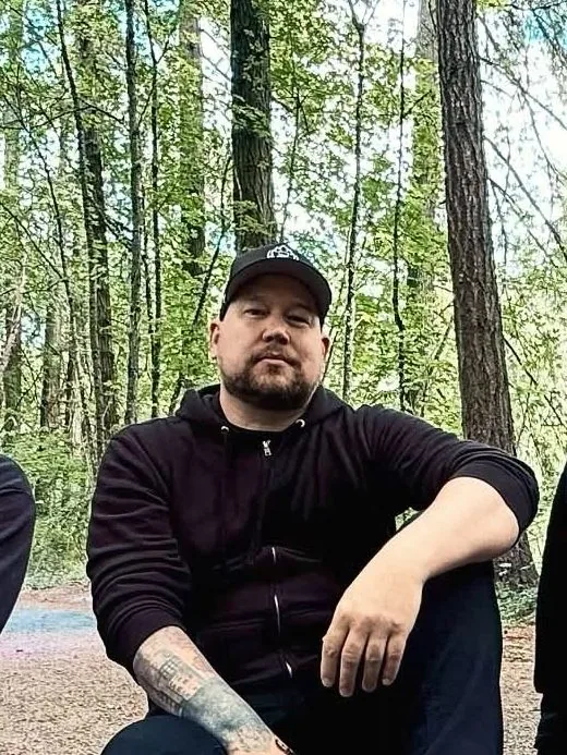 Graham Brown in a black cap and hoodie with tattooed forearms, sitting in the forest