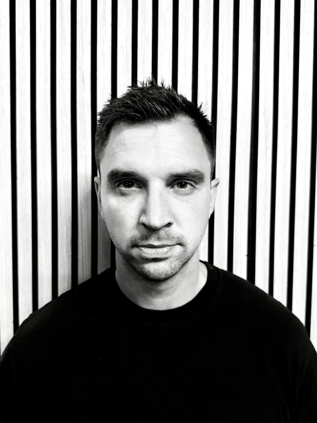 Kramer, with short dark hair and a black tee, in front of a slatted wall