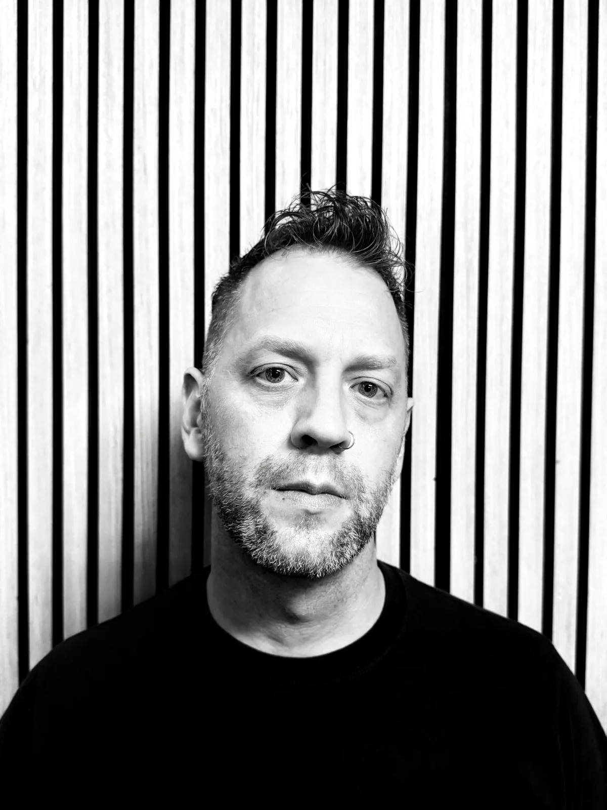 Michael, with a nose ring and black tee, in front of a slatted wall